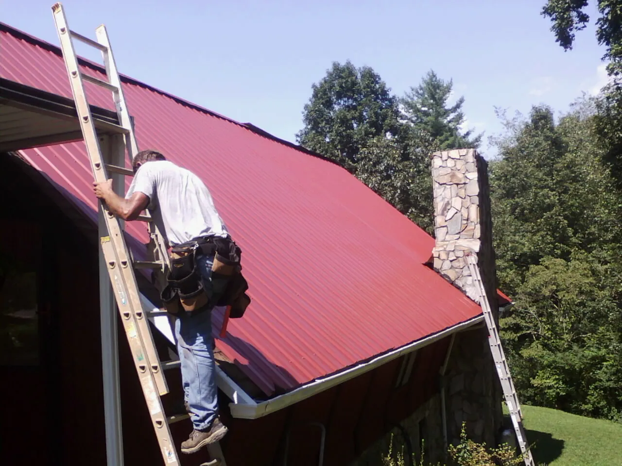 Roofer on a ladder beside a red metal roof and stone chimney — Soffit & Fascia Repair by Roofing Brothers in Mount Carroll